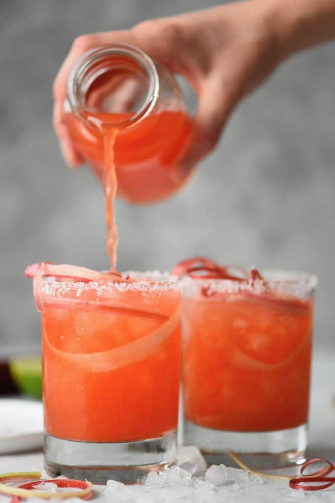 Pouring raspberry rhubarb margarita into glass with rhubarb ribbons