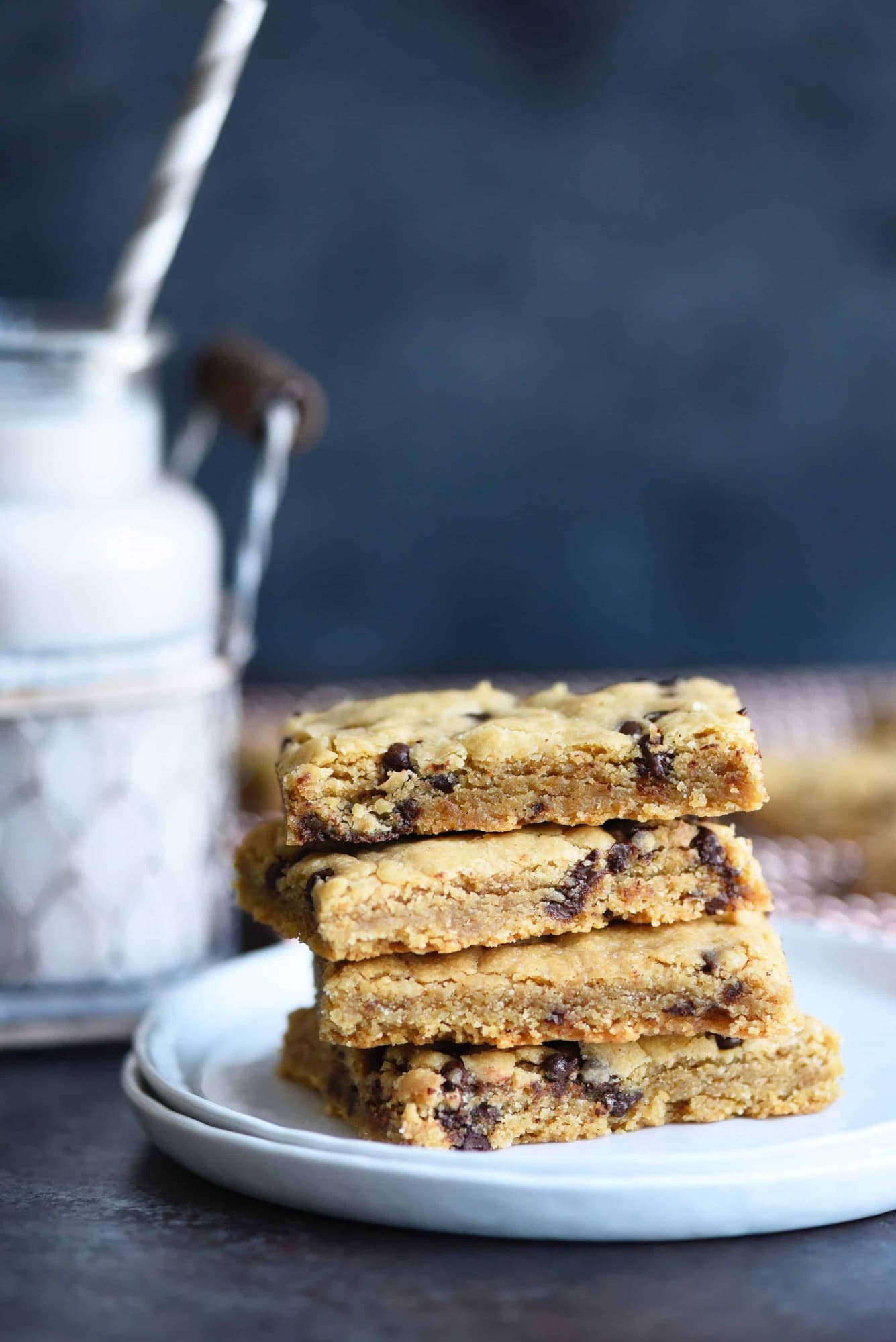 Side view of stack of chocolate chip cookie bars on a white plate with milk in background