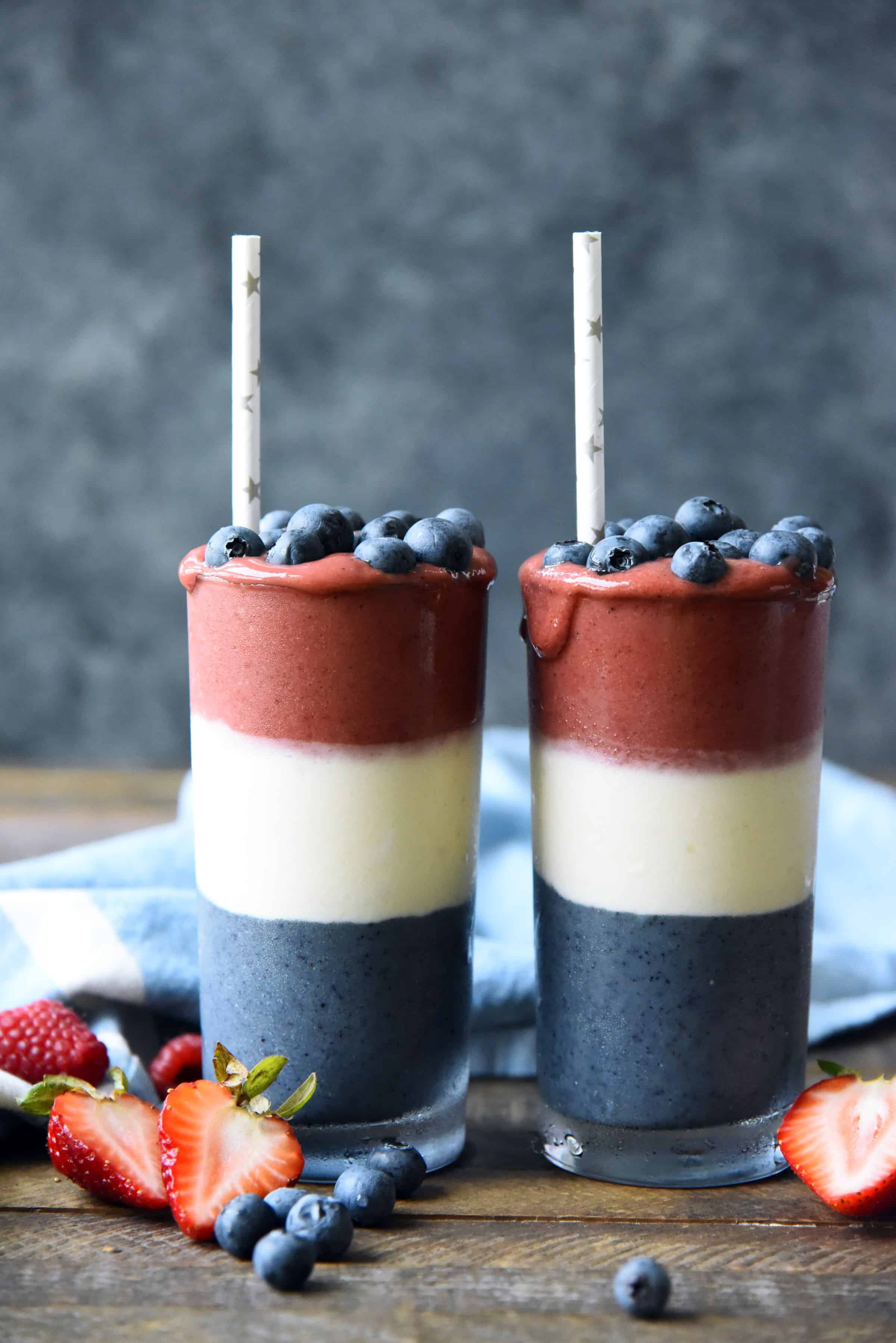 Side view of red white and blue layered smoothies topped with blueberries on a wood background