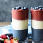 Side view of red white and blue layered smoothies topped with blueberries on a wood background