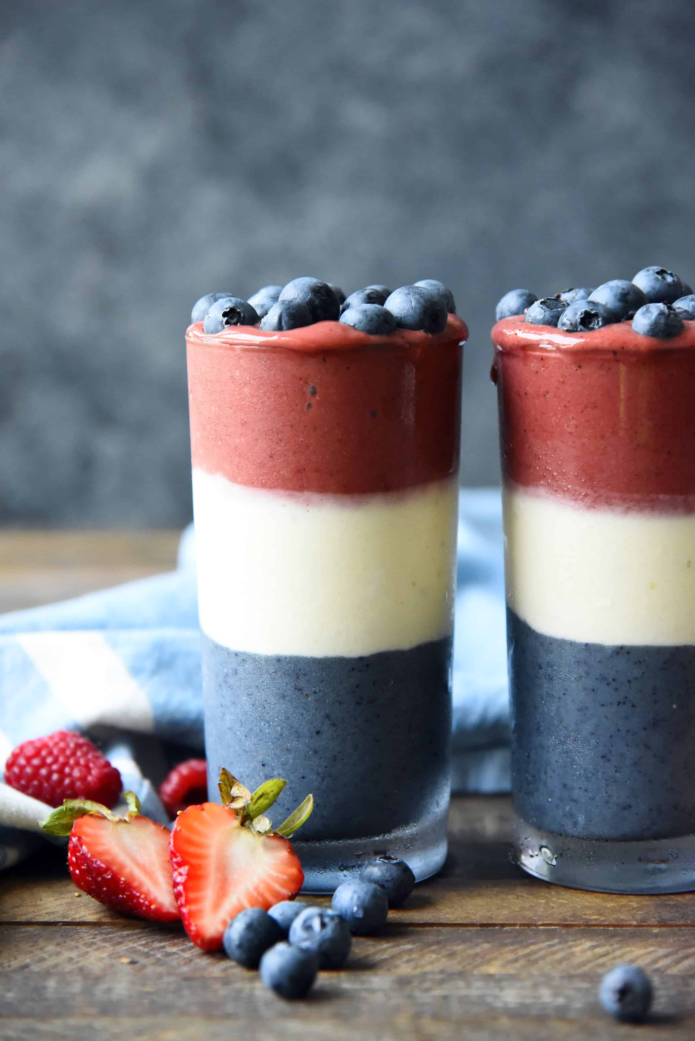Side view of red white and blue layered smoothies topped with blueberries on a wood background