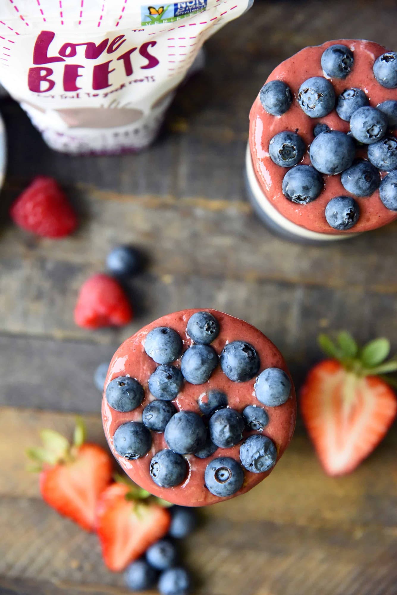 Overhead of red white and blue layered smoothies topped with blueberries on a wood background