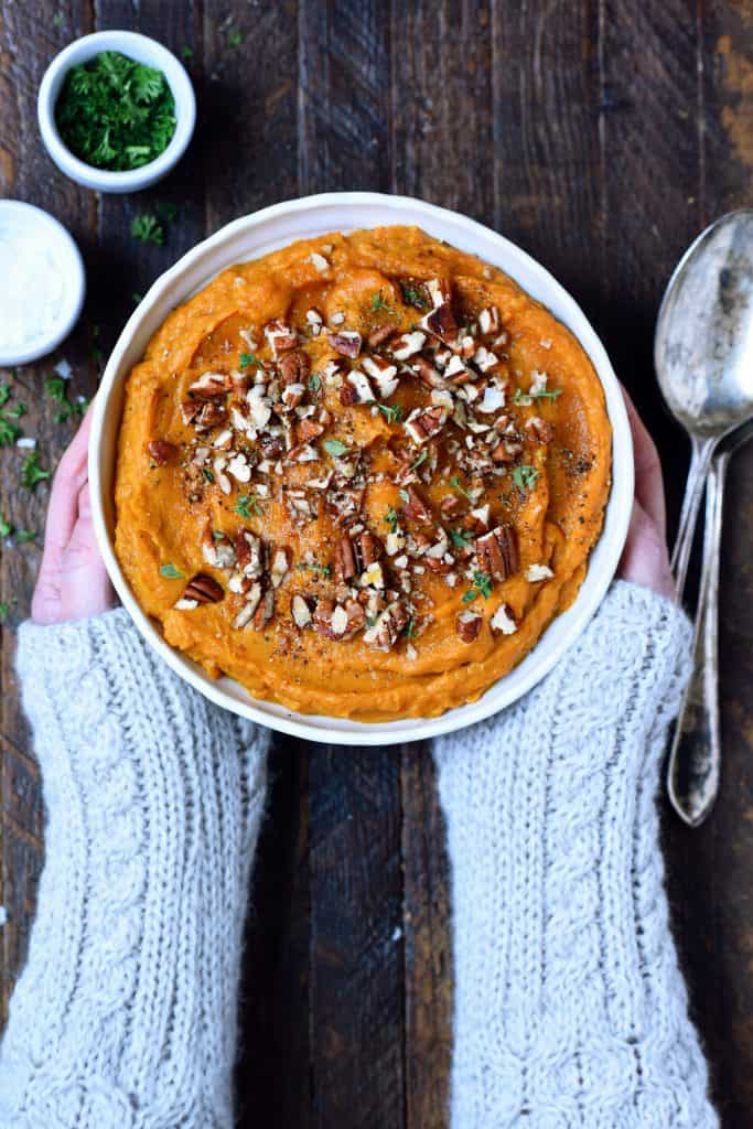 Person in gray sweater holding bowl of mashed sweet potatoes with chopped pecans