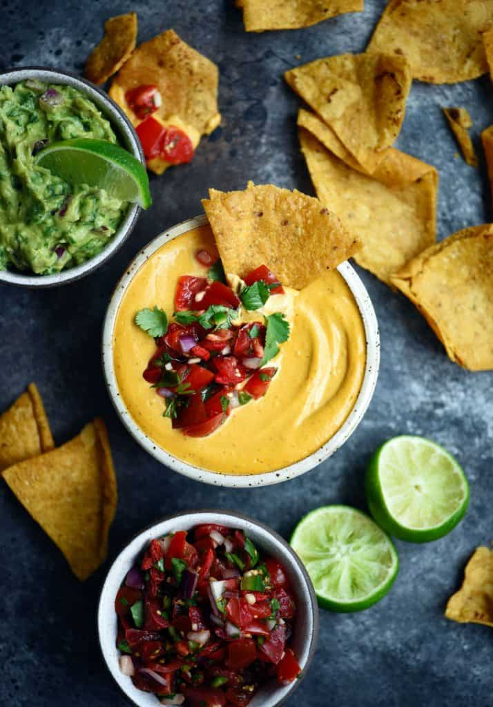Vegan queso in a small white bowl with salsa, guacamole and tortilla chips