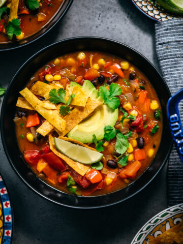 overhead view of vegan tortilla soup in black bowls topped with avocado, tortilla strips and lime.