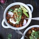 Overhead of vegan chili in a white bowl topped with avocado, lime, crispy lentils