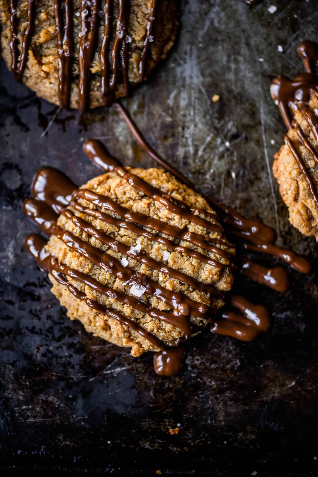 Overhead of peanut butter cookie drizzled with dark chocolate and salt on a dark, rustic cookie sheet