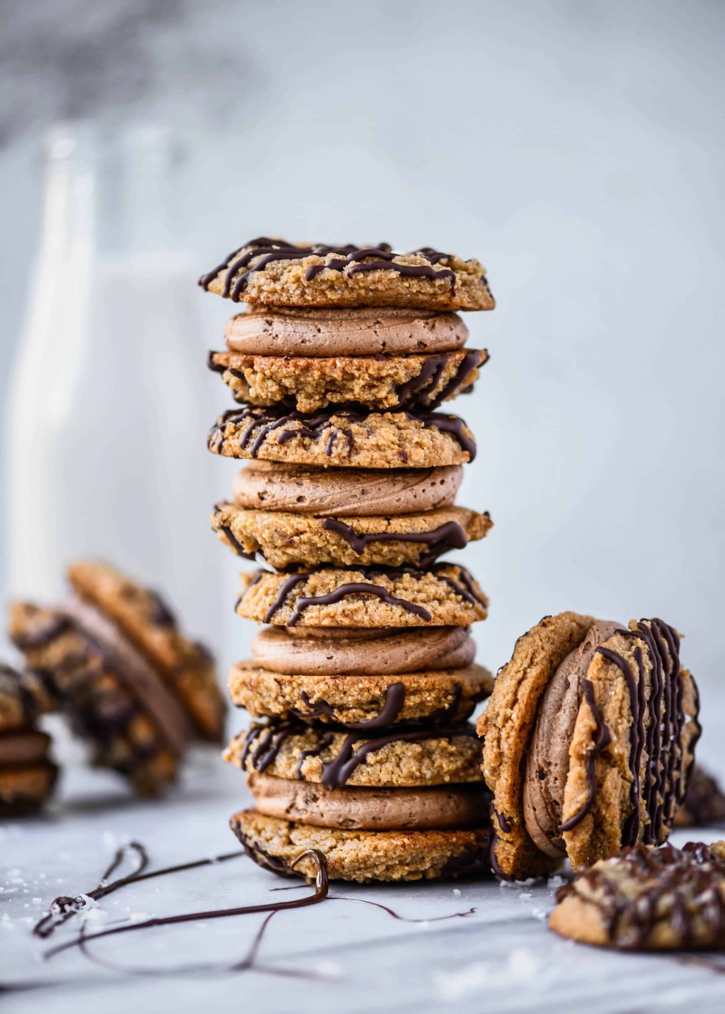Stack of peanut butter sandwich cookies drizzled with dark chocolate on a white marble background with jug of milk