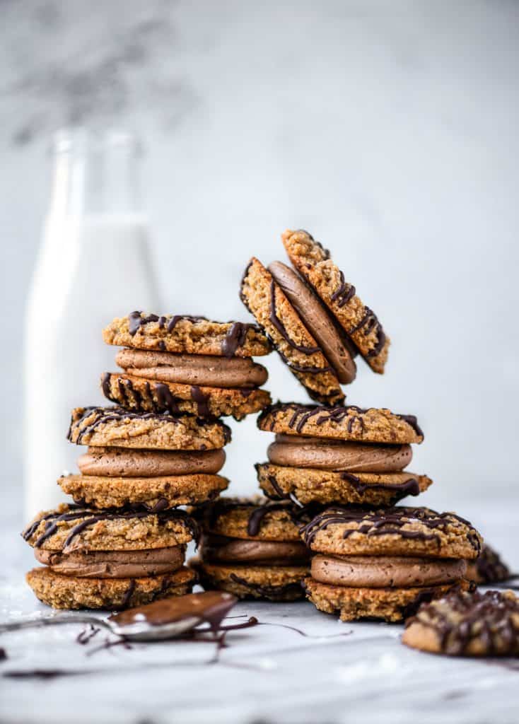 Stack of peanut butter sandwich cookies drizzled with dark chocolate on a white marble background with jug of milk