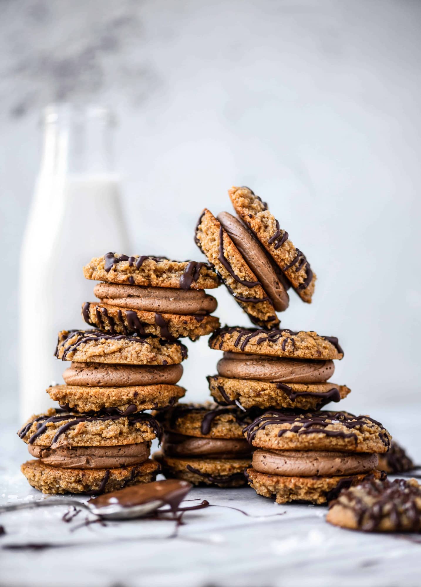 Stack of peanut butter sandwich cookies drizzled with dark chocolate on a white marble background with jug of milk