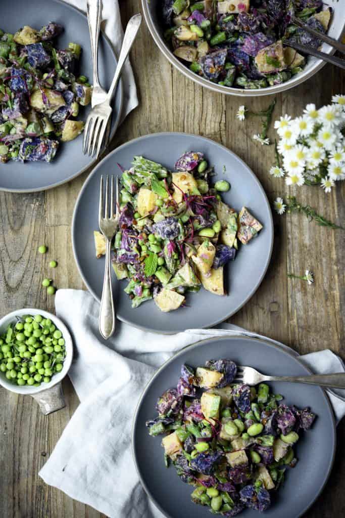 Overhead of purple potato spring salad in a white bowl with microgreens and fresh peas on a gray plate with an antique fork