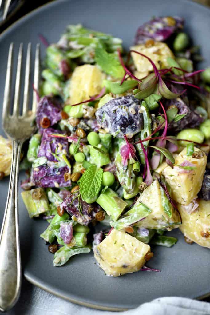 Close up of purple potato spring salad in a white bowl with microgreens and fresh peas on a gray plate with an antique fork