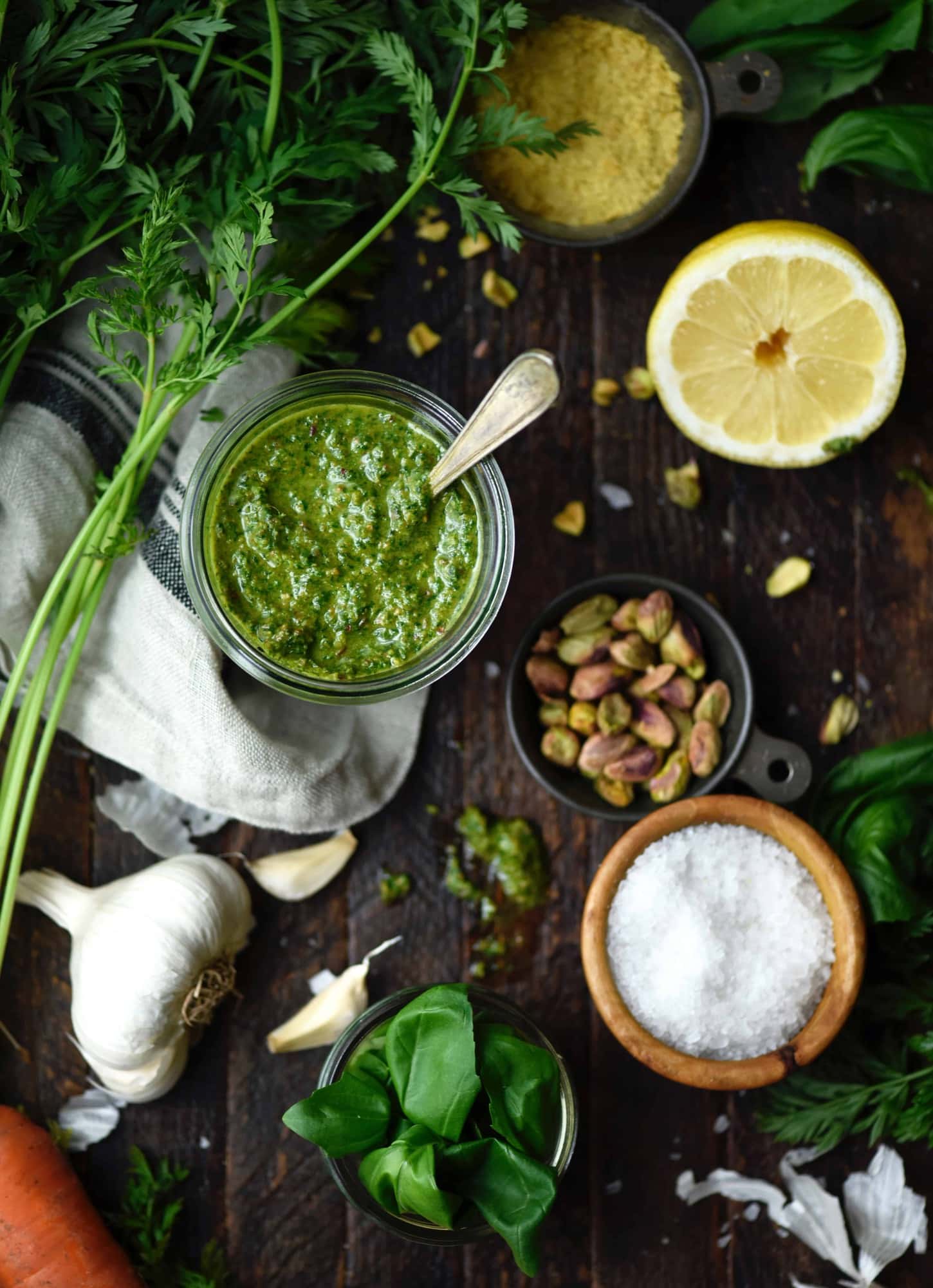 Overhead of homemade pesto in a jar surrounded by ingredients including lemon, pistachios, salt, a head of garlic, carrot tops and fresh basil