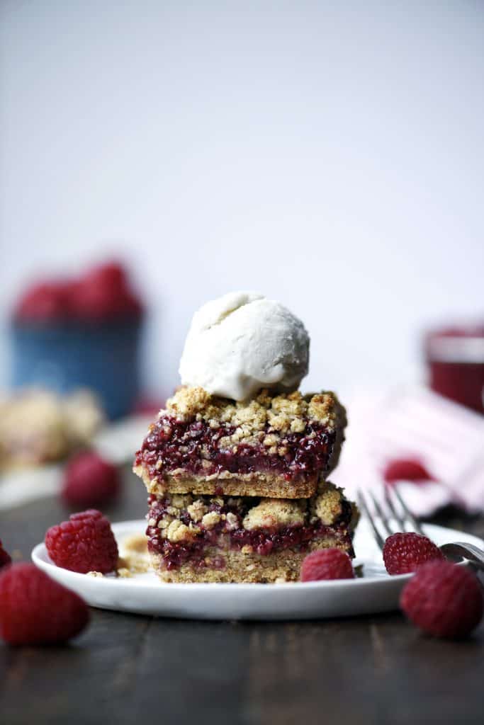 Stacked gluten free raspberry oatmeal crumble bars with a scoop of vanilla ice cream and fresh raspberries on a small white plate
