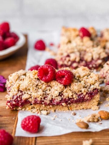 close up side view of raspberry crumble bar topped with fresh raspberries on parchment paper.