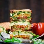Side view of vegan caprese and avocado sandwich sliced in half on a wood board with fresh heirloom tomatoes