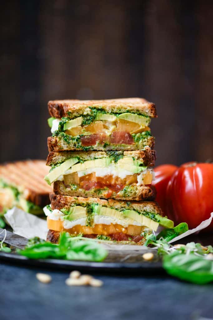Side view of vegan caprese and avocado sandwich sliced in half on a wood board with fresh heirloom tomatoes