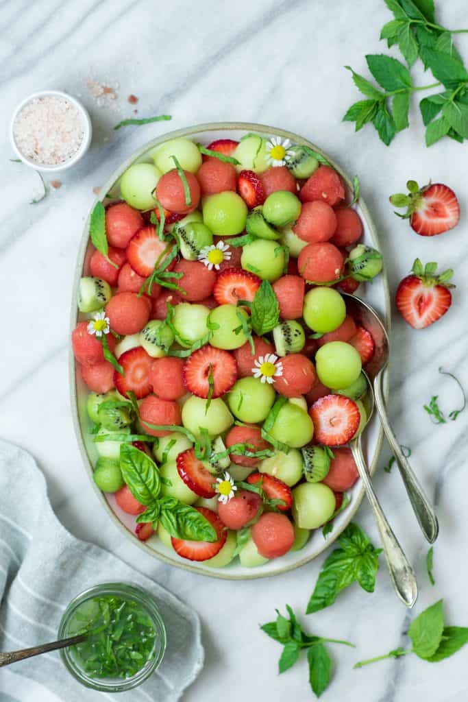 Overhead of melon ball salad on a white platter with strawberries and a lime mint dressing on a white marble backdrop