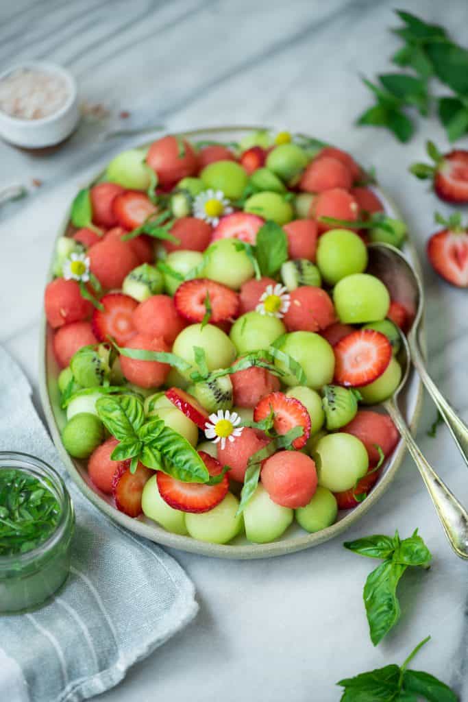 Side view of melon ball salad on a white platter with strawberries and a lime mint dressing on a white marble backdrop