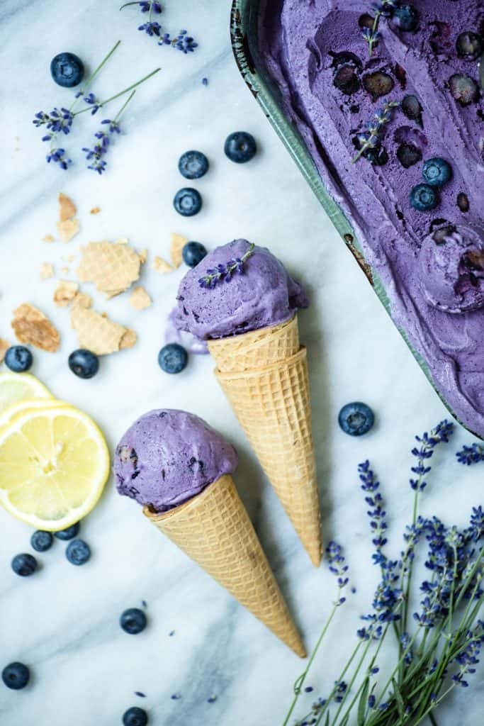 Two cones of blueberry lavender vegan ice cream on a white marble background with blueberries and lavender