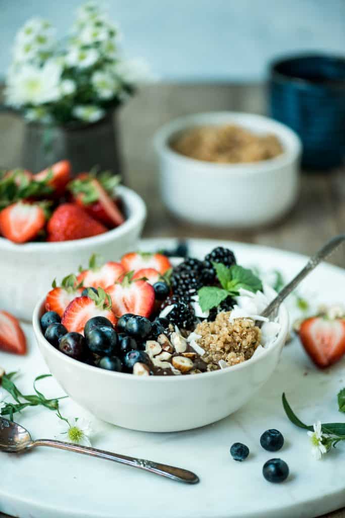 Side view of breakfast quinoa in a white bowl with fresh berries, coconut and hazelnuts on a white marble serving tray