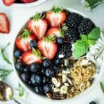 Close up overhead of breakfast quinoa in a white bowl with fresh berries, coconut and hazelnuts on a white marble serving tray