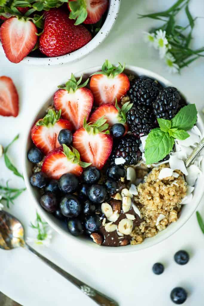 Close up overhead of breakfast quinoa in a white bowl with fresh berries, coconut and hazelnuts on a white marble serving tray