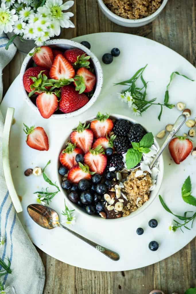 Overhead of breakfast quinoa in a white bowl with fresh berries, coconut and hazelnuts on a white marble serving tray