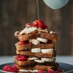 Maple syrup pouring over a stack of french toast with sliced bananas and raspberries