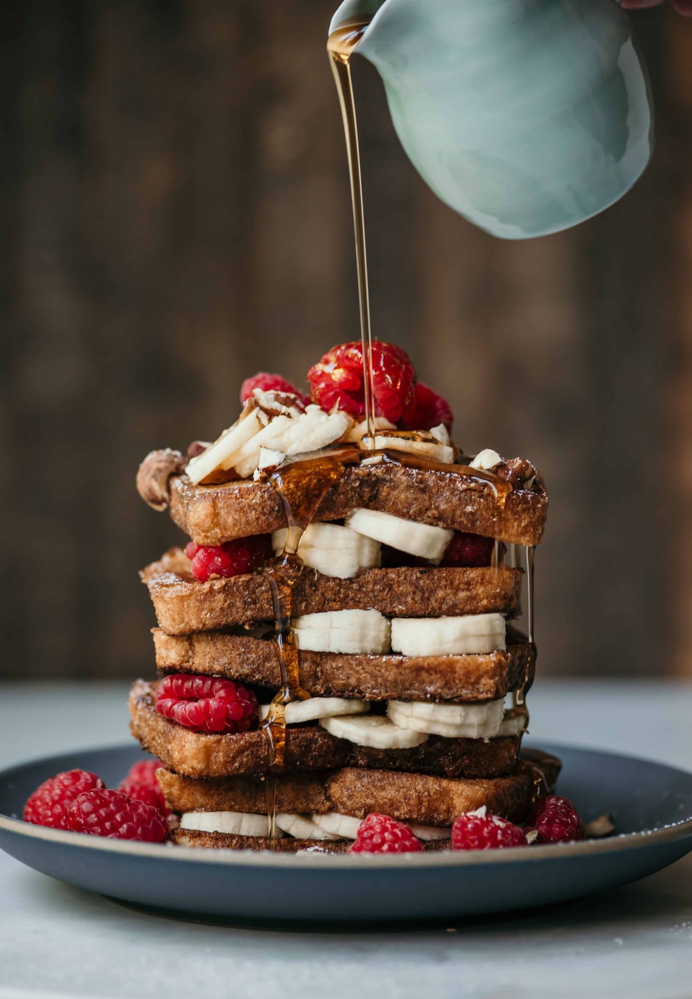 Maple syrup pouring over a stack of french toast with sliced bananas and raspberries