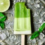 Close up of green popsicle with bit taken out on a sheet pan with crushed ice