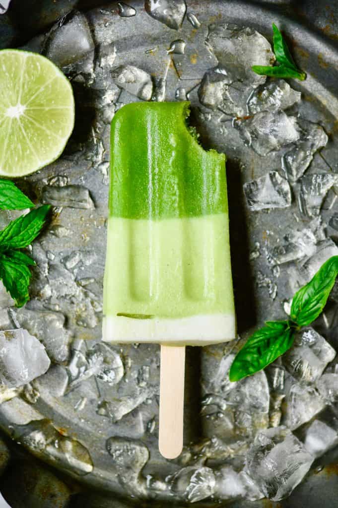 Close up of green popsicle with bit taken out on a sheet pan with crushed ice