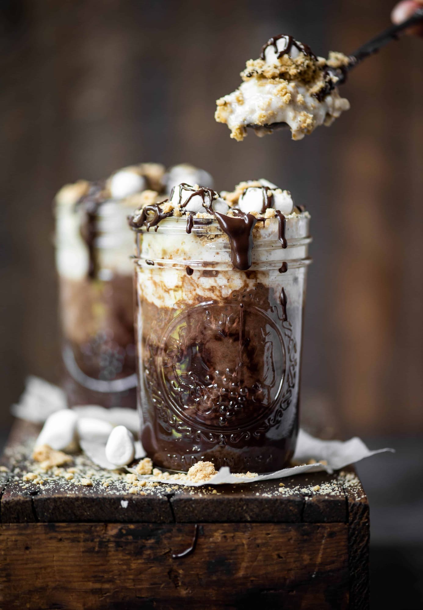 Spoon taking bite out of s'mores overnight oats drizzled with chocolate and marshmallows on a rustic wood board