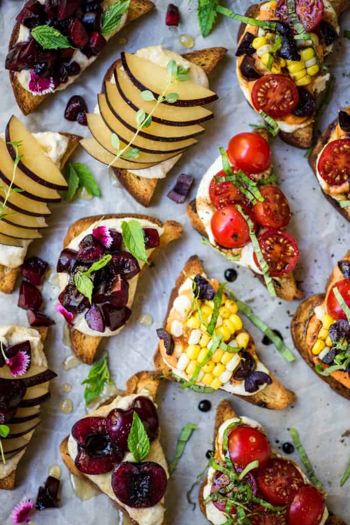 Overhead of crostini topped with ricotta, herbs, tomatoes, cherries and plums
