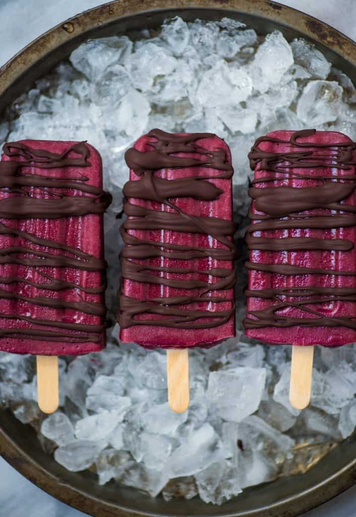 Overhead of three red popsicles drizzled with chocolate over crushed ice on a tray
