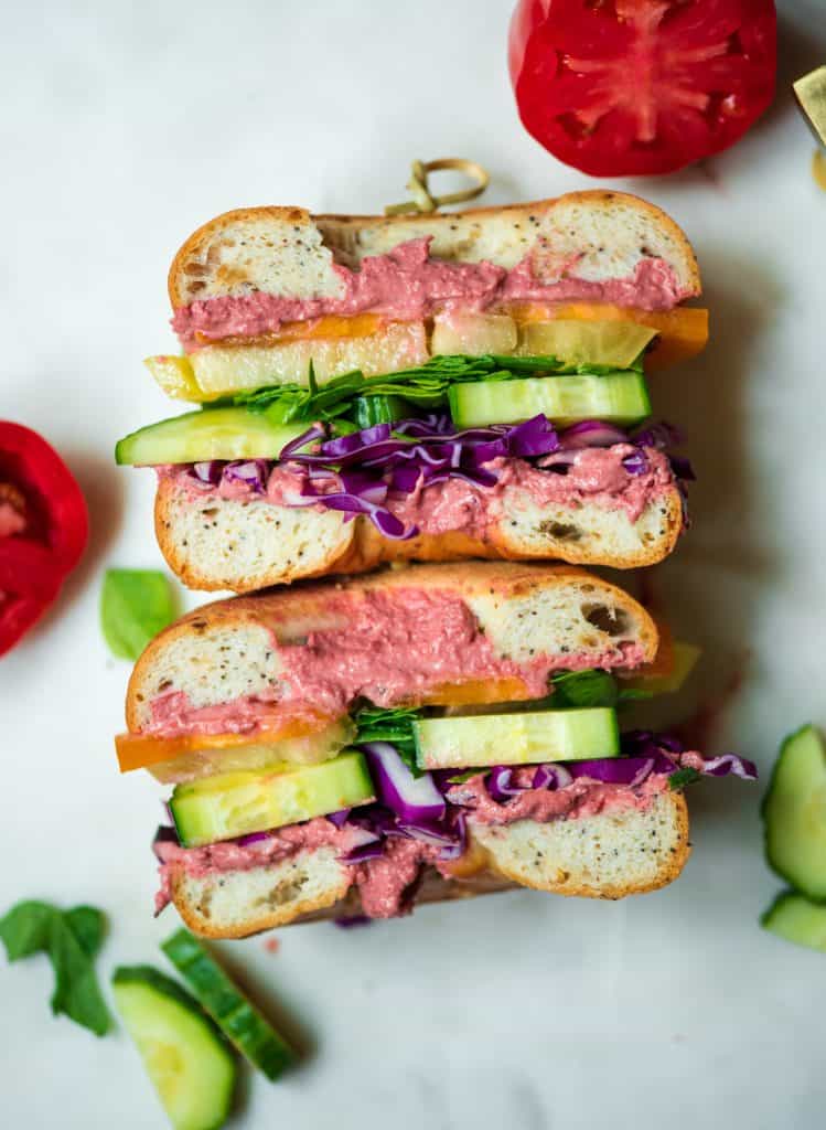 Overhead of rainbow veggie bagel sandwich with beet hummus sliced in half on a white tray