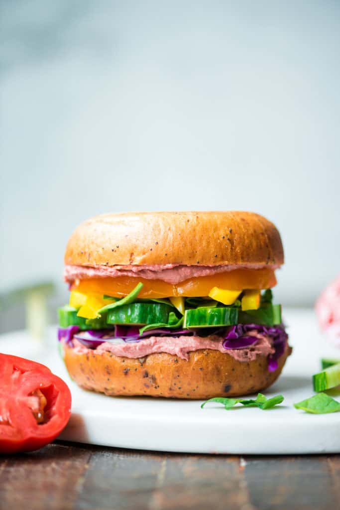 Side view of rainbow vegetable bagel sandwich with beet hummus on a white serving tray