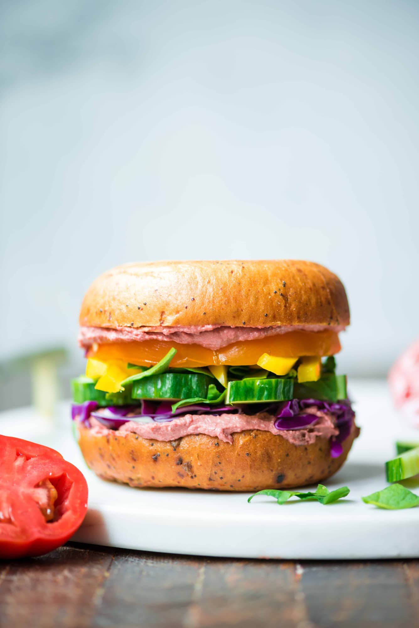 Side view of rainbow vegetable bagel sandwich with beet hummus on a white serving tray