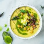 Overhead of yellow tomato and pepper soup topped with herb oil corn and chickpeas in a white bowl on a white marble surface