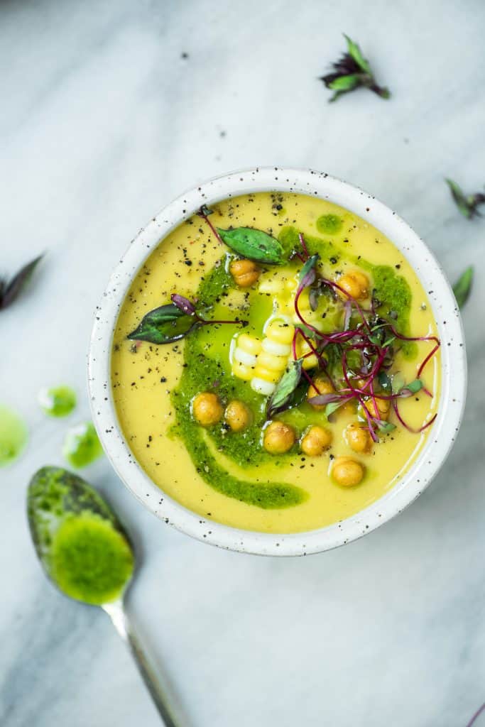 Overhead of yellow tomato and pepper soup topped with herb oil corn and chickpeas in a white bowl on a white marble surface