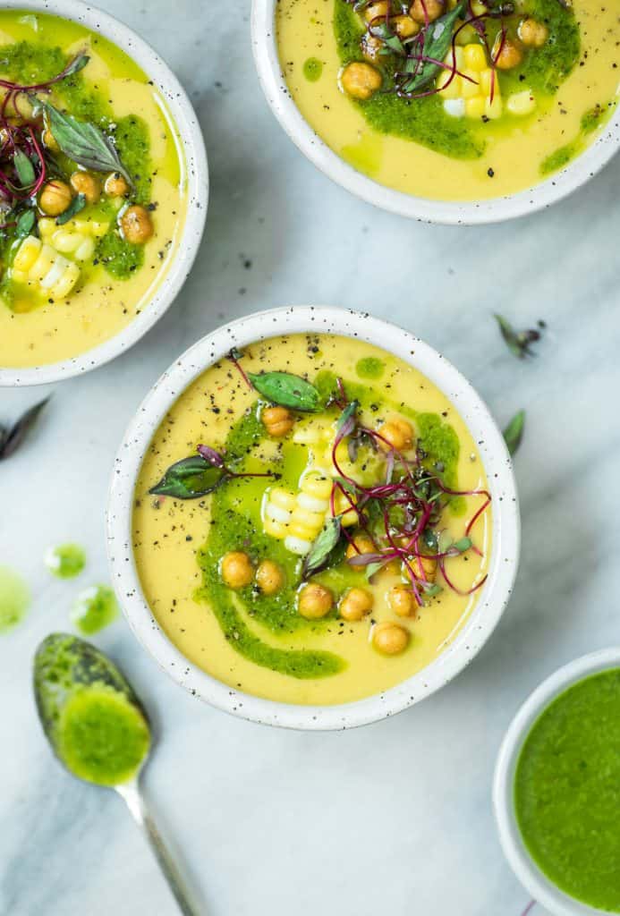 Overhead of 3 bowls of yellow tomato and pepper soup topped with herb oil corn and chickpeas on a white marble surface