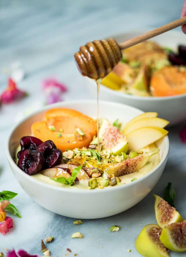 Drizzling honey onto yogurt bowl topped with fresh fruit and pistachios
