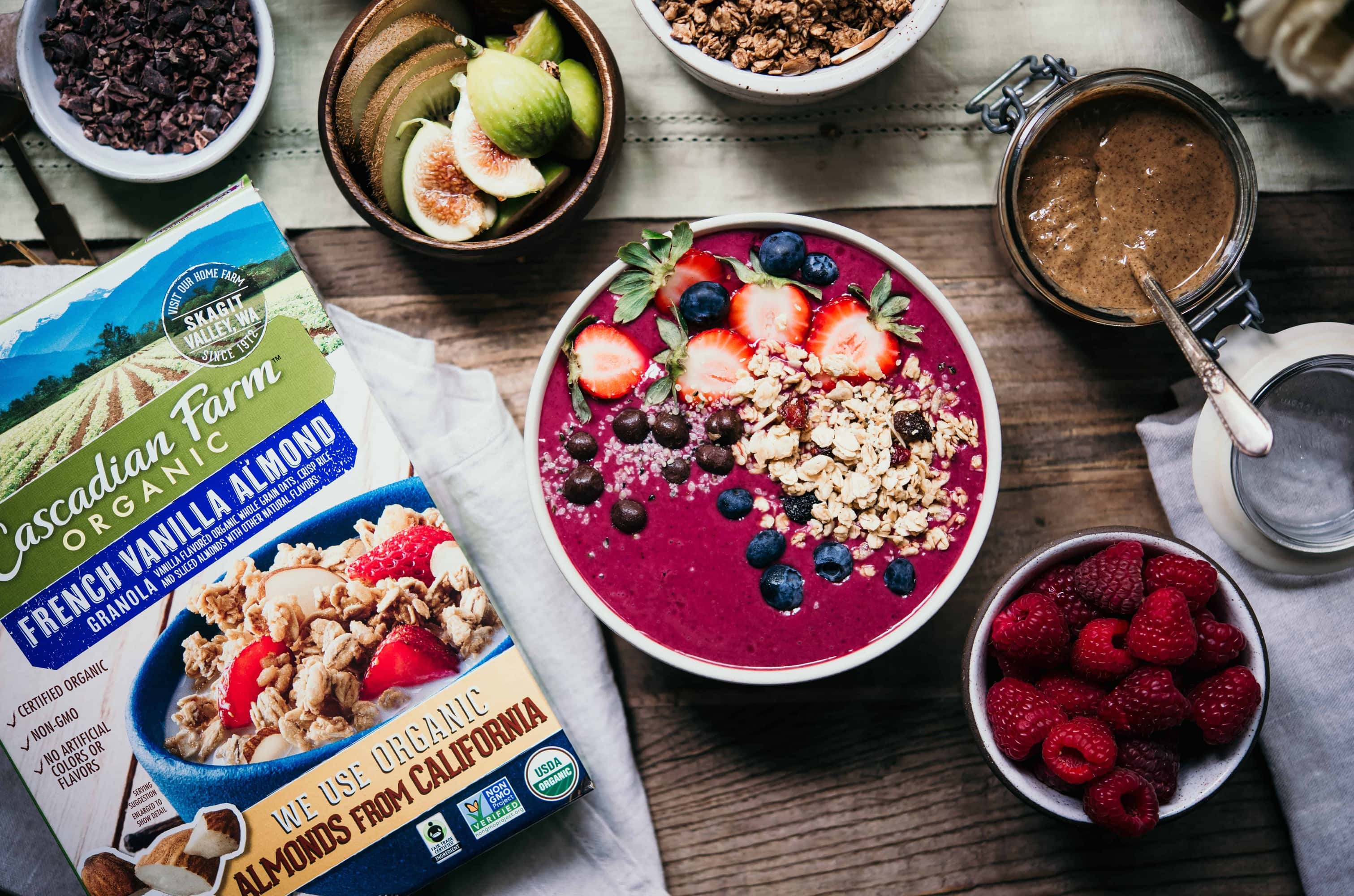 Pink Smoothie Bowl topped with fresh fruit and granola on a table with fresh flowers, bowls of fruit and granola