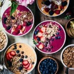Four pink and orange smoothie bowls topped with fresh berries and granola on a wood table with fresh flowers