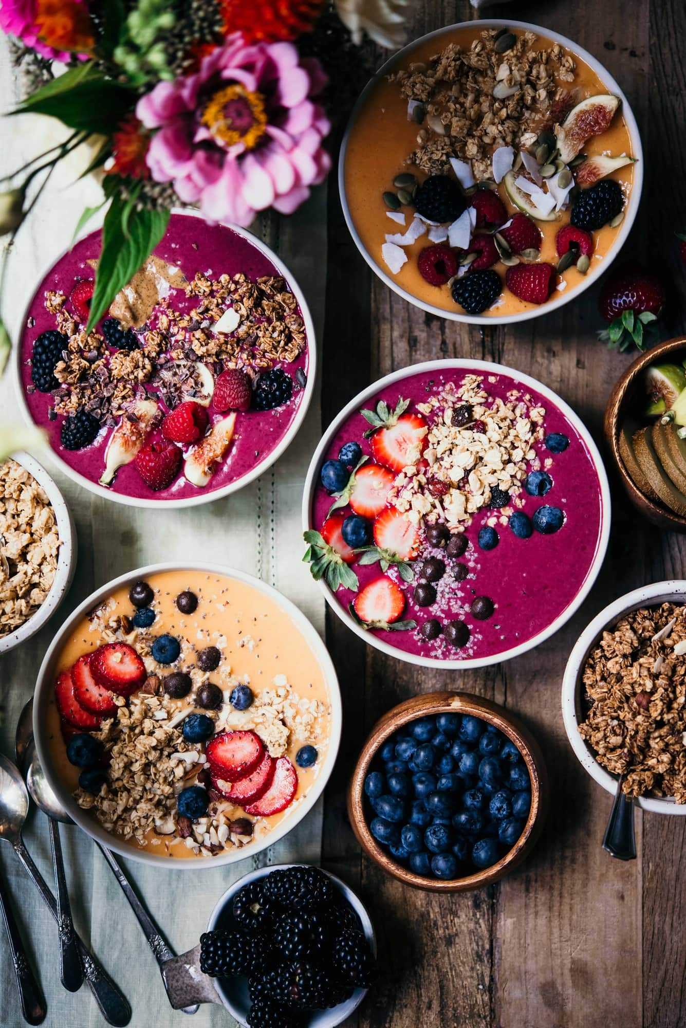 Four pink and orange smoothie bowls topped with fresh berries and granola on a wood table with fresh flowers