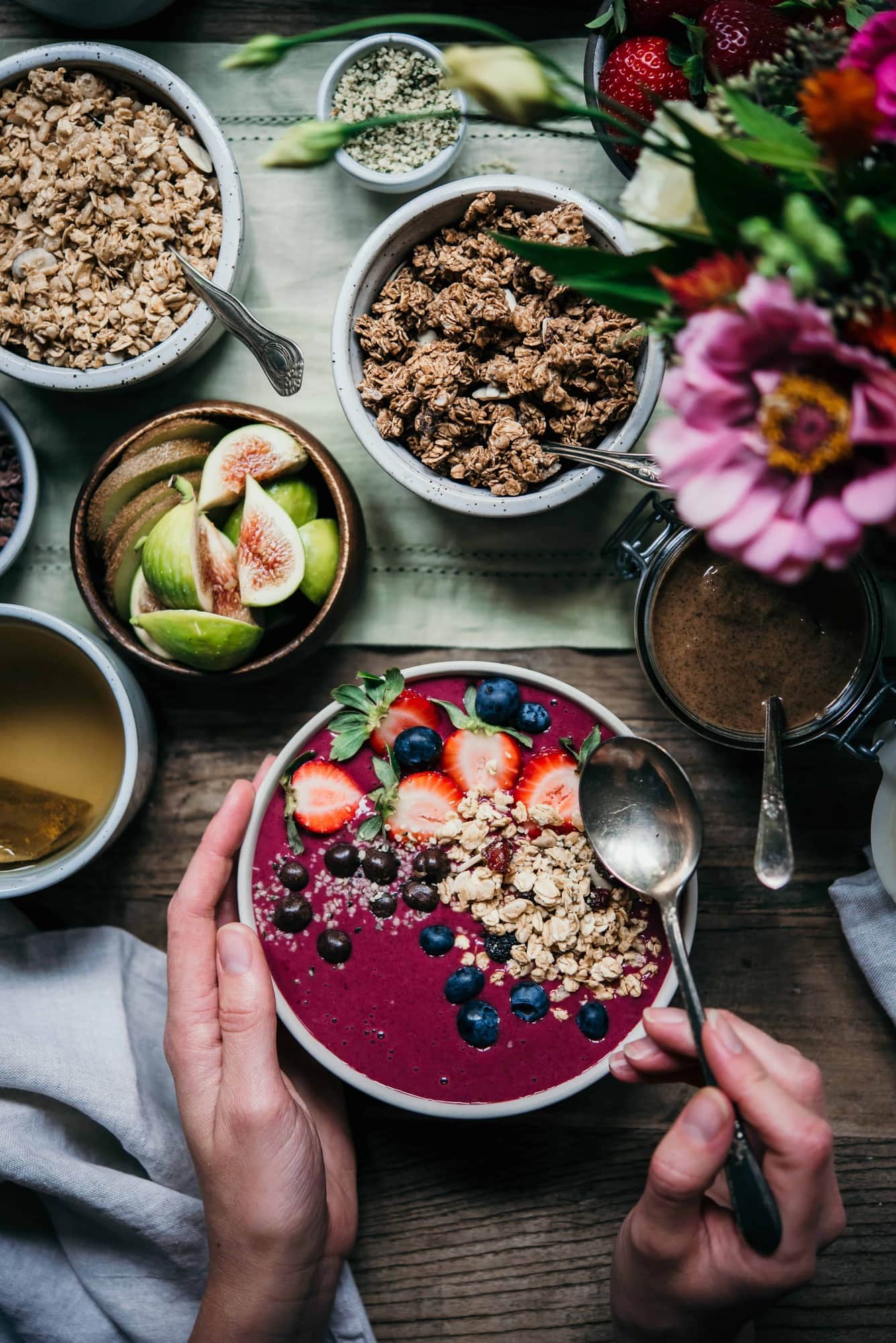 Pink Smoothie Bowl topped with fresh fruit and granola on a table with fresh flowers, bowls of fruit and granola