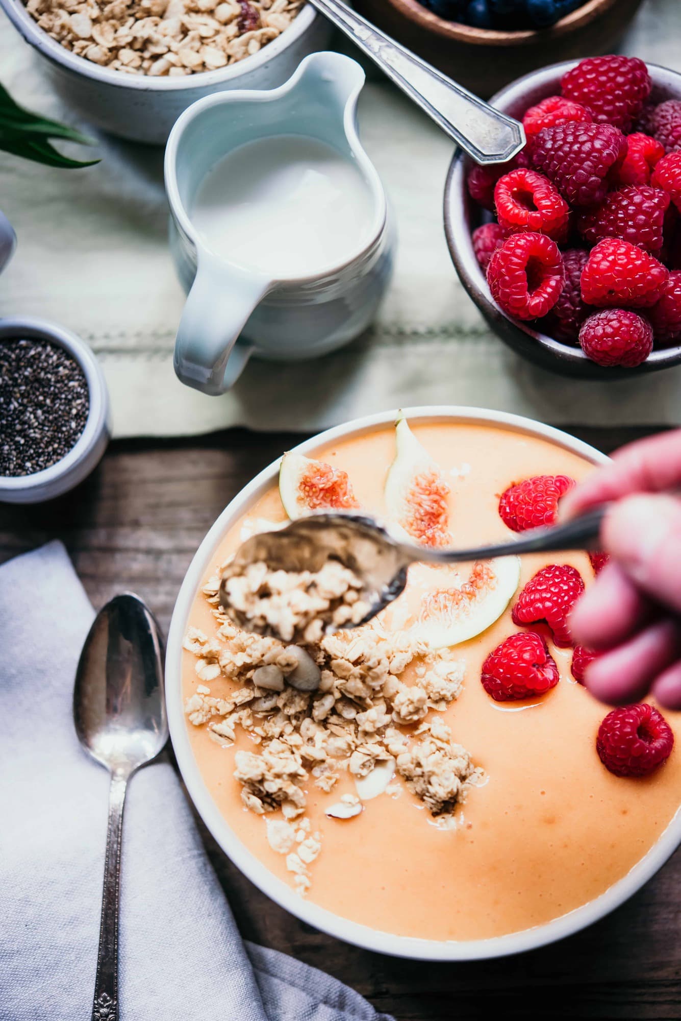 Overhead of hand pouring granola onto an orange smoothie bowl topped with fresh raspberries