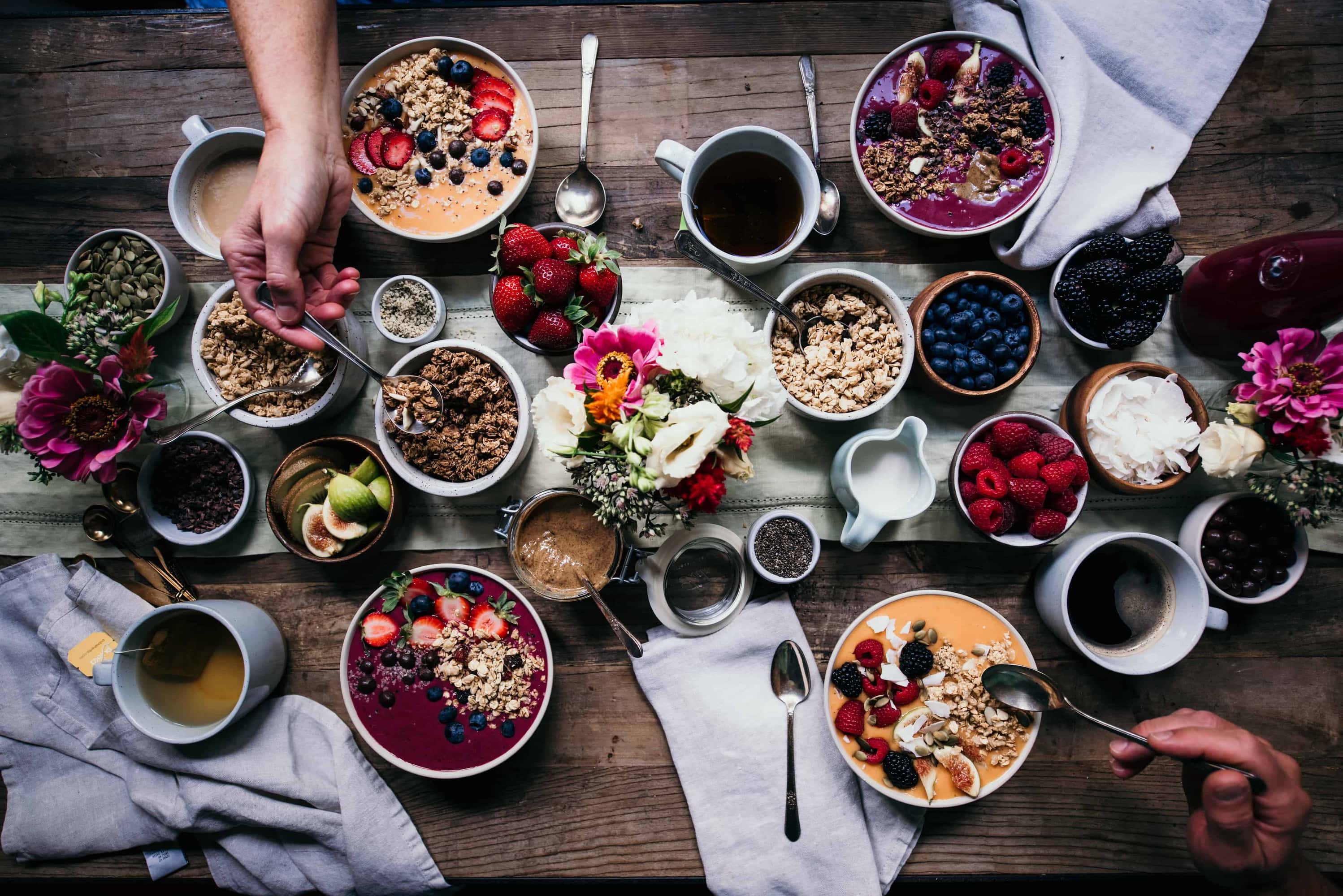 Overhead of breakfast tablescape with smoothie bowls, bowls of fresh fruit, granola, flowers, tea, coffee