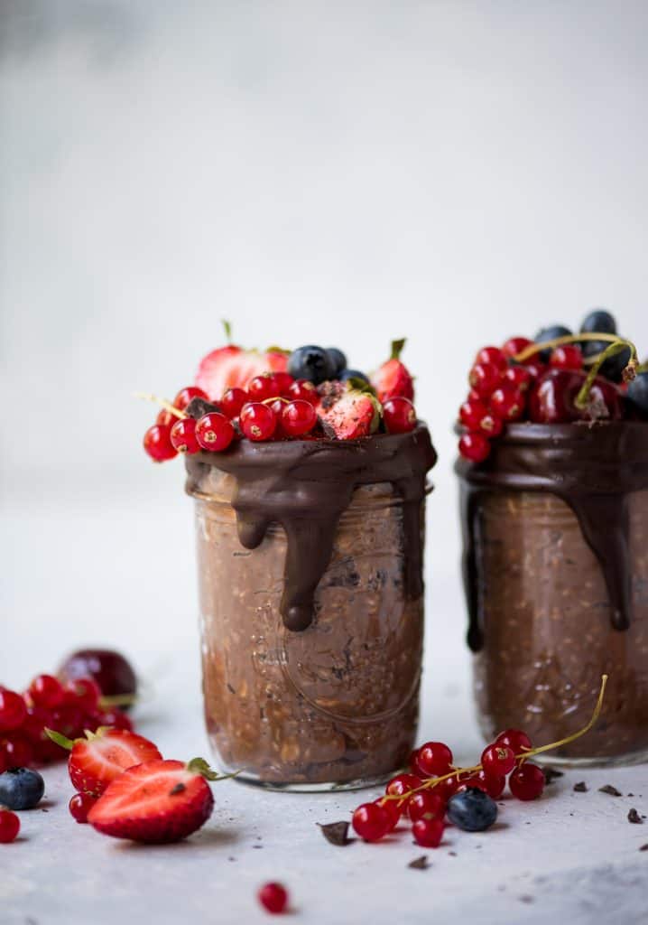 Side view of chocolate mocha overnight oats in a glass jar with dark chocolate drizzle and fresh summer berries. Styled on a white background
