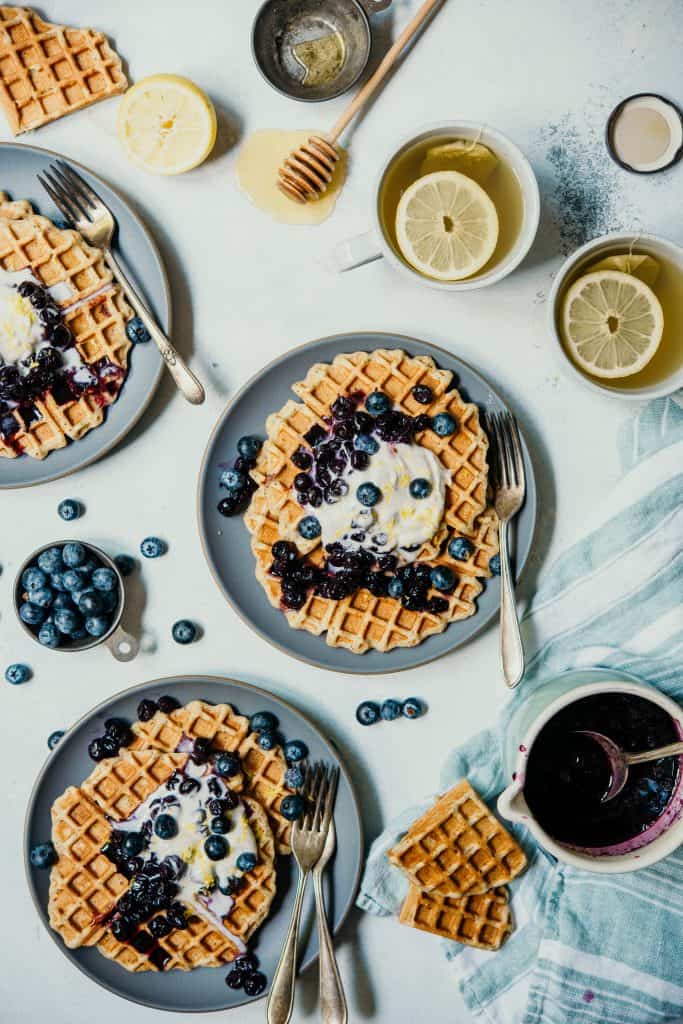 Overhead of 3 plates of lemon blueberry poppyseed waffles topped with whipped cream and blueberries on a white background